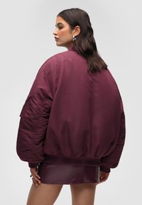 Burgunderröd bomberjacka med ledig passform, ribbade muddar och en slät yta. Har en ficka på vänster ärm.