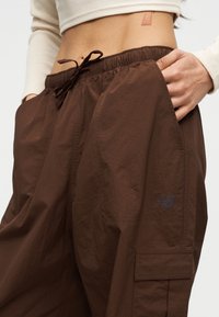 Άτομο που φορά καφέ cargo παντελόνι με κορδόνι στη μέση και χέρι στην τσέπη, συνδυασμένο με κοντό μπεζ μακρυμάνικο τοπ.