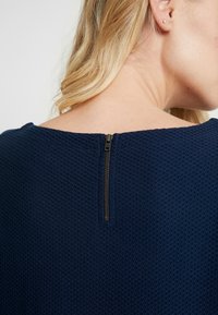 Haut en tissu texturé navy avec encolure bateau et fermeture éclair courte à l'arrière. Gros plan sur l'encolure avec des détails soulignant la fermeture éclair.