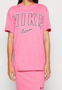 Rosa bomullströja med svart "NIKE" grafik och logotyp. Avslappnad passform, korta ärmar, rund halsringning. Slät textur, minimalistisk design.