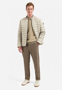 Lichtbeige pufferjack met een geribbelde trui eronder, bruine broek en witte sneakers. Zachte textuur, horizontale stiksels en ritssluiting.