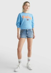 Felpa azzurra con ricamo "Tommy Jeans", abbinata a pantaloni shorts in denim. Sneakers bianche e calze con logo completano il look.