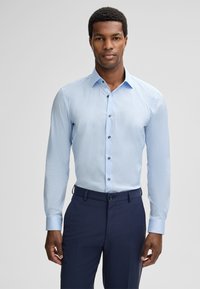 Man in een lichtblauw overhemd met knopen en een donkerblauwe broek, staand tegen een effen lichtgrijze achtergrond.