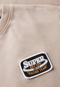 Beige sweatshirtstof med ribbet krave. Har et sort og hvidt broderet mærke med teksten "Super Dry JPN" og "Racing Team" i orange.