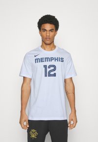 T-shirt blanc Nike avec "MEMPHIS" en texte bleu et le numéro "12" en dessous, à manches courtes, col rond, porté avec un pantalon noir à motif jaune.