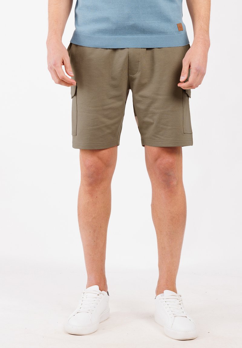 Pantaloni cargo in tessuto verde oliva, dotati di tasche frontali e tasche cargo laterali. Abbinati a un maglione blu e scarpe da ginnastica bianche.