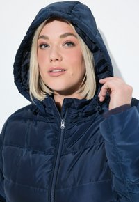 Chaqueta de plumas azul marino con capucha ajustable, cierre de cremallera y diseño acolchado. Presenta una textura suave y brillante, y puños elásticos.
