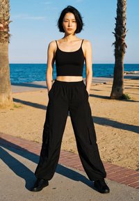 Young woman in black crop top, cargo pants, and sneakers stands between two palm trees near a beach with blue sky and sea in the background.