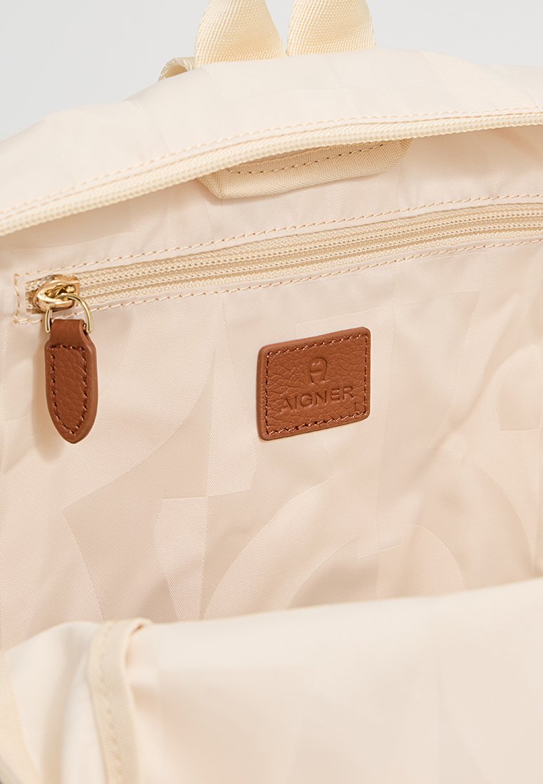 Beige Rucksackinnenseite mit einem Reißverschlussfach, glattem Stoff und einem braunen Lederlogopatch. Verfügt über ein strukturiertes Muster im Futter.