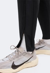 Μαύρο παντελόνι με πλαϊνό φερμουάρ μισάνοιχτο στον αστράγαλο πάνω από λευκές κάλτσες, σε συνδυασμό με μπεζ παπούτσια τρεξίματος Nike ZoomX με μαύρο λογότυπο Swoosh.