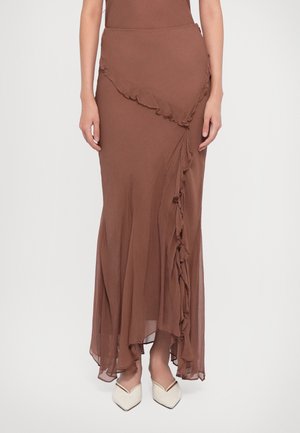 Jupe longue asymétrique marron avec des détails à volants portée avec des chaussures blanches à bout pointu, montrée de la taille jusqu'aux pieds.
