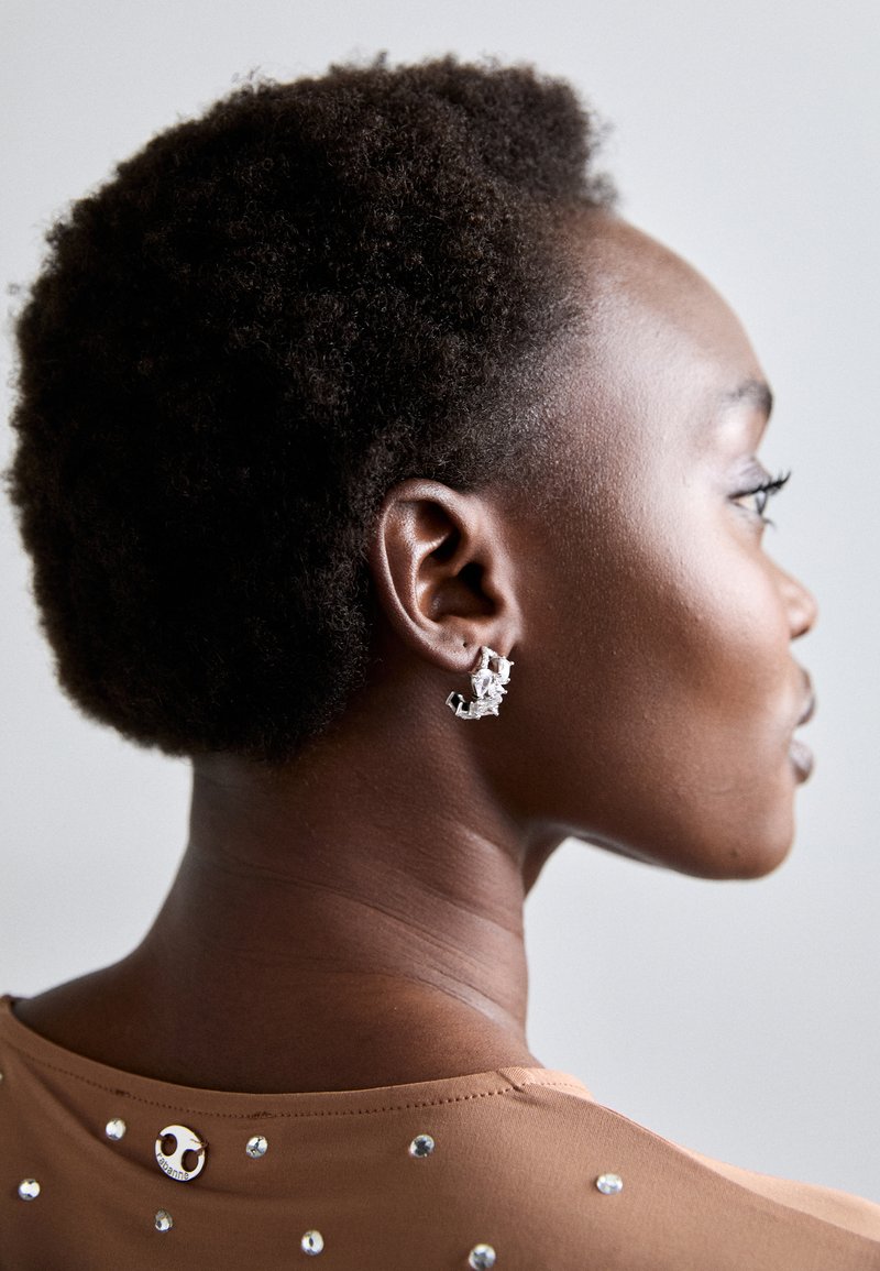 Ασημένια γεωμετρικά σκουλαρίκια με ανάγλυφη σχεδίαση ορατά στο αυτί. Λείο, κοντό φυσικό μαλλί και τοπ σε ταμπά με λεπτομέρειες από παγιέτες στο πίσω μέρος.