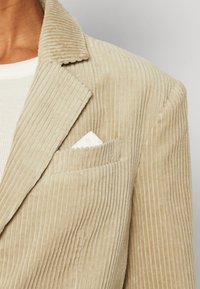 Beige fløjl blazer med fremtrædende lodrette ribber. Har skrå krave og et hvidt lommetørklæde synligt i det venstre brystlomme.