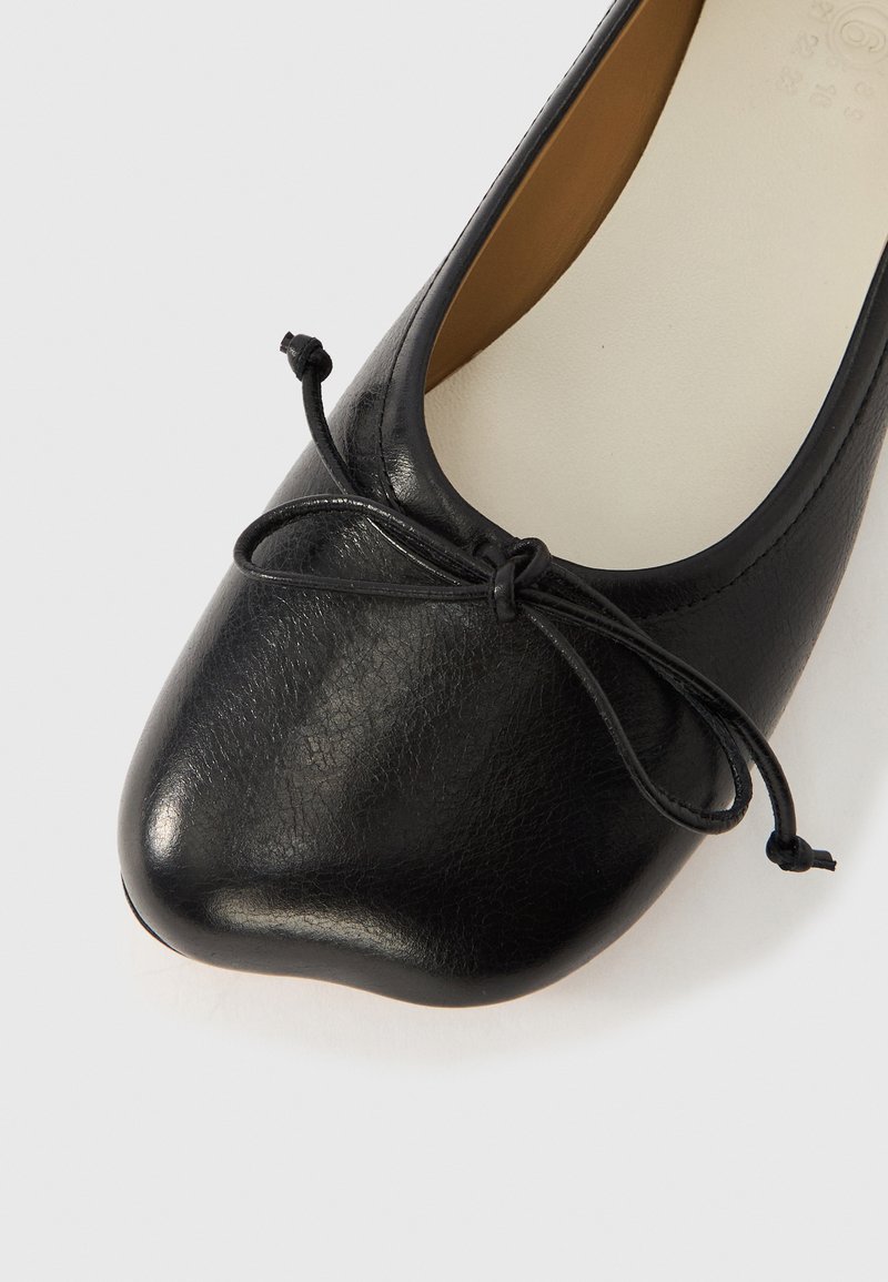 Zapato plano de cuero negro con punta redondeada, superficie texturizada y un detalle de lazo en la pala. Plantilla de color crema visible en el interior.