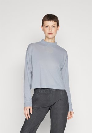 Top court à manches longues bleu clair avec un col montant et un logo ton sur ton sur la poitrine, associé à un pantalon gris foncé.