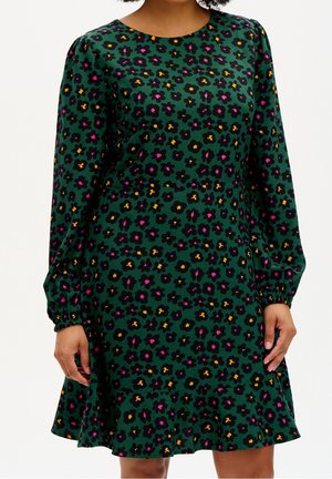 Groene jurk met lange mouwen, bedrukt met roze, gele en zwarte bloemen en een geribbelde zoom, gedragen door een persoon met ontspannen handen langs de zij.