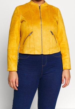 Veste zippée en suède jaune avec deux poches avant zippées portée avec un jean bleu foncé sur une personne devant un fond blanc.