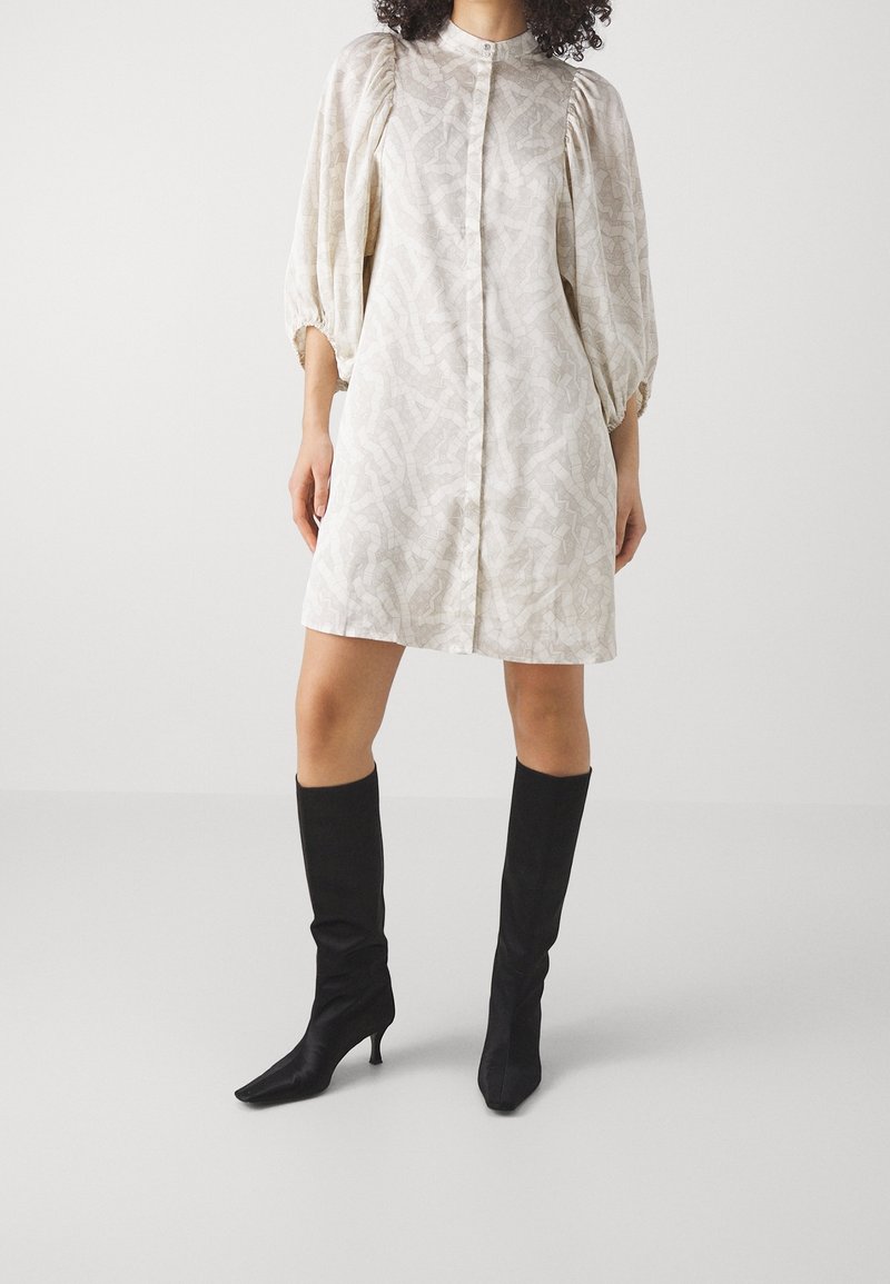Vestito beige chiaro, svasato e morbido con maniche a sbuffo, motivo testurizzato e chiusura a bottoni; abbinato a stivali neri al ginocchio con tacco.