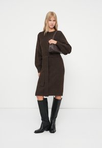 Donna che indossa un vestito a maglia belted marrone scuro, stivali neri fino al ginocchio e tiene una piccola borsa scura sulla spalla, in piedi su uno sfondo bianco.