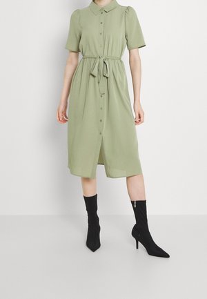 Femme portant une robe midi boutonnée vert clair avec des manches courtes et une ceinture à nouer, associée à des bottines noires à bout pointu avec des fermetures éclair.