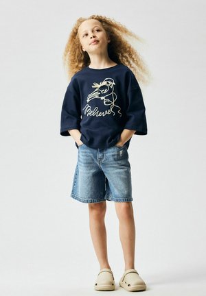 Dijete s kovrčavom kosom, odjeveno u tamnoplavu majicu s pticom i natpisom "Believer", traper kratke hlače i bež cipele bez vezica, stoji s rukama u džepovima.