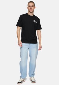 Camiseta de algodón negra con un logo blanco en el lado izquierdo del pecho, combinada con unos jeans holgados de color azul claro y zapatillas en blanco y negro.