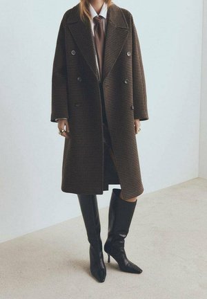 Personne portant un manteau croisé marron à carreaux, une chemise blanche, une cravate marron et des bottes noires montantes à talons, debout à l’intérieur sur un sol beige.