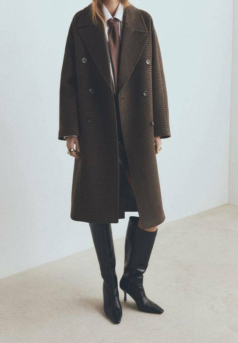 Personne portant un manteau croisé marron à carreaux, une chemise blanche, une cravate marron et des bottes noires montantes à talons, debout à l’intérieur sur un sol beige.