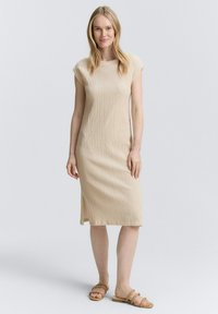 Abito lavorato a maglia beige con silhouette dritta, maniche a cappuccio e spacco laterale. Realizzato in tessuto testurizzato, lunghezza al ginocchio con una morbida caduta.