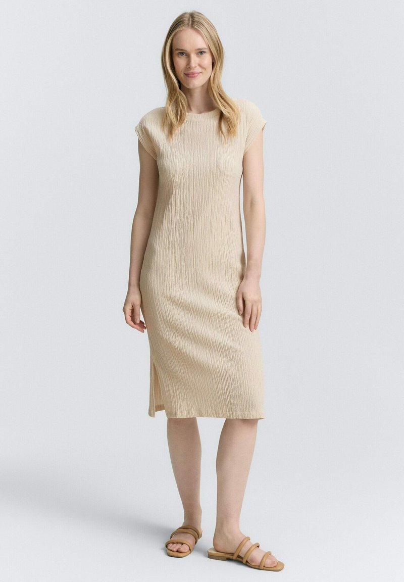 Abito lavorato a maglia beige con silhouette dritta, maniche a cappuccio e spacco laterale. Realizzato in tessuto testurizzato, lunghezza al ginocchio con una morbida caduta.