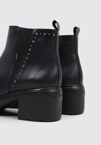 Botas de tobillo de cuero negro con tacón cuadrado, con lados elásticos y remaches plateados decorativos a lo largo de la costura exterior. Suela texturizada.