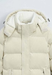 Veste en doudoune en velours côtelé crème avec doublure en polaire douce, col haut et deux poches latérales, présentant une texture à côtes verticales.