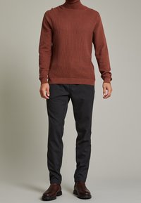 Pull à col roulé côtelé marron à manches longues, associé à un pantalon rayé foncé. Le mannequin porte des bottes en cuir marron.