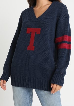 Kvinde iført en mørkeblå strikket V-hals sweater med en stor rød "T" foran og to røde striber på den ene ærme.