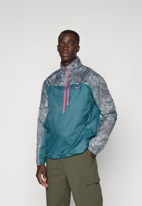 Teal halv-zip jakke med et mønstret grått overparti, rosa glidelås og elastiske ermer; har en logo-patch på brystet, laget av lett materiale.