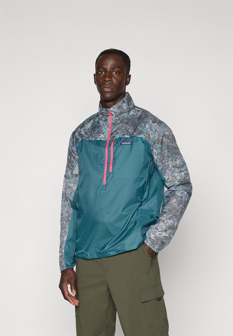 Teal halv-zip jakke med et mønstret grått overparti, rosa glidelås og elastiske ermer; har en logo-patch på brystet, laget av lett materiale.