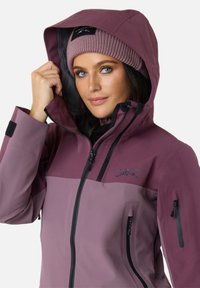 Purple two-tone jacket with a zip front, adjustable hood, and multiple pockets. A knitted pink beanie complements the look.