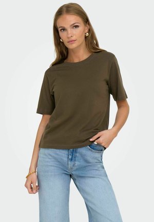 Giovane donna che indossa una maglietta a maniche corte verde oliva e jeans azzurri, posando con una mano in tasca su uno sfondo semplice.