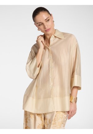 Frau trägt locker sitzendes beiges Satinkleid mit Knöpfen und Dreiviertelärmeln, florale gemusterte Hose, goldenes Armband und Creolen.