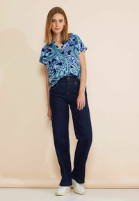 Camicia azzurra a maniche corte con motivo paisley e dettagli verdi; abbinata a jeans scuri a vita alta e sneaker bianche.