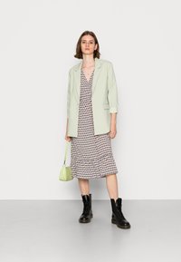 Blazer verde-claro sobre um vestido estampado com bainha em babados. Botas pretas com atacadores e uma pequena bolsa verde completam o look.