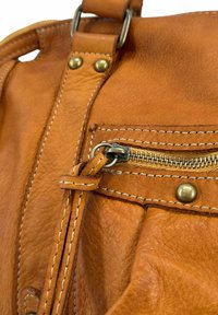 Gros plan d'un sac en cuir beige avec des coutures détaillées, une fermeture éclair en métal, des rivets et une sangle enroulée autour d'un anneau en métal.