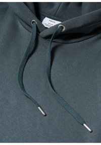 Sweat à capuche bleu sarcelle foncé avec cordons passés dans des œillets métalliques et embouts métalliques, affichant une étiquette blanche indiquant la taille à l'intérieur du col.
