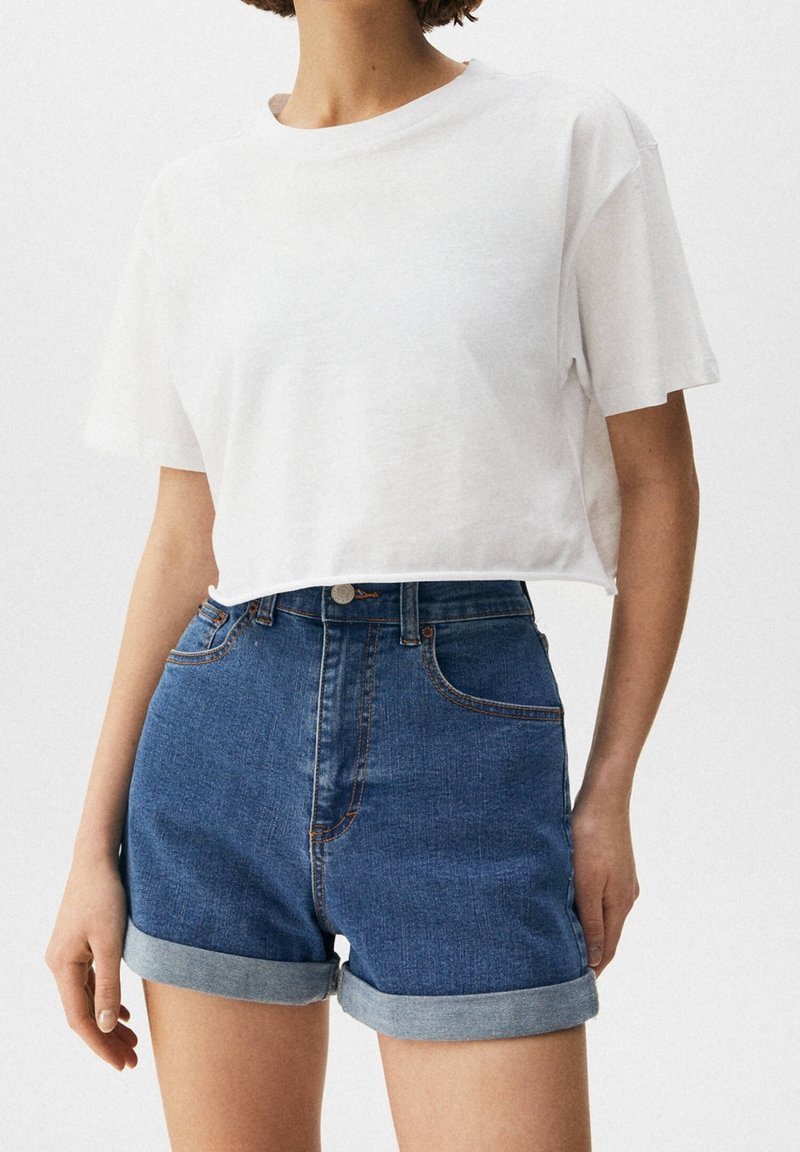 Wit cropped T-shirt met een ronde halslijn, gecombineerd met blauw denim shorts met een hoge taille, omgeslagen zomen en klassieke metalen accenten.