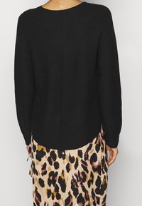 Pull noir en maille avec un col rond et des manches longues, présentant un ourlet plus long à l'arrière. Porté avec une jupe à motif léopard.