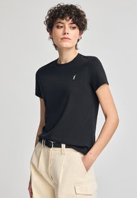 T-shirt nera in cotone con scollo a girocollo e maniche corte, con un piccolo logo bianco sul petto. Abbinata a pantaloni chiari.