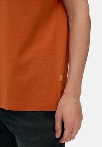 Orange bomulds T-shirt med korte ærmer, der har en sømkant detalje og et lille gult mærke tæt på kanten. Sammen med mørke jeans.