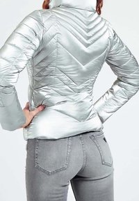 Sølv pufferjakke med en skinnende finish, der har et chevron-quiltet mønster og en høj krave. Båret med grå jeans.