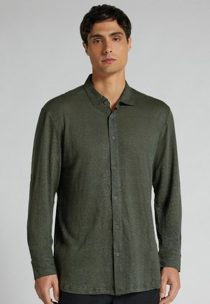 Mann, der ein langärmeliges, olivgrünes Hemd mit Knöpfen trägt, steht vor einem schlichten hellgrauen Hintergrund.