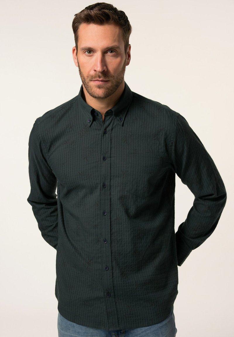 Camisa abotonada verde oscuro hecha de una tela texturizada con sutiles patrones de hojas, con cuello y mangas largas, combinada con jeans de mezclilla.